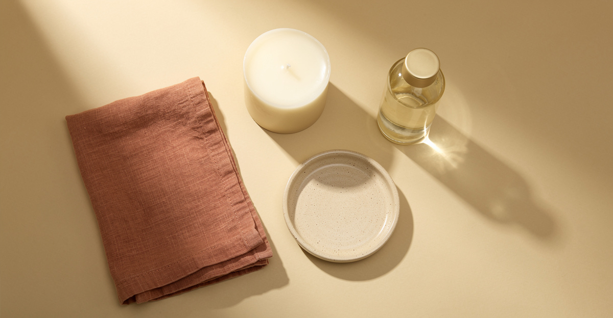 An overhead arrangement of a folded textile, candle, ceramic dish and glass bottle on a warm cream surface, representing product category selection