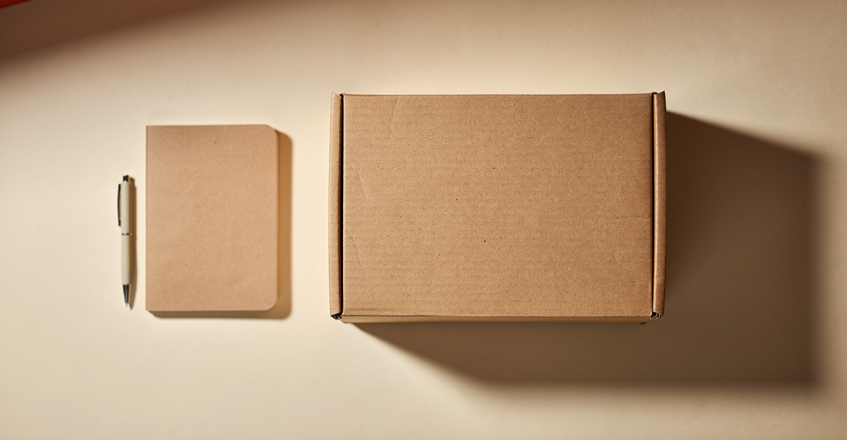 A single kraft shipping box beside a notebook and pen on a warm cream desk, representing the first steps of setting up a dropshipping store