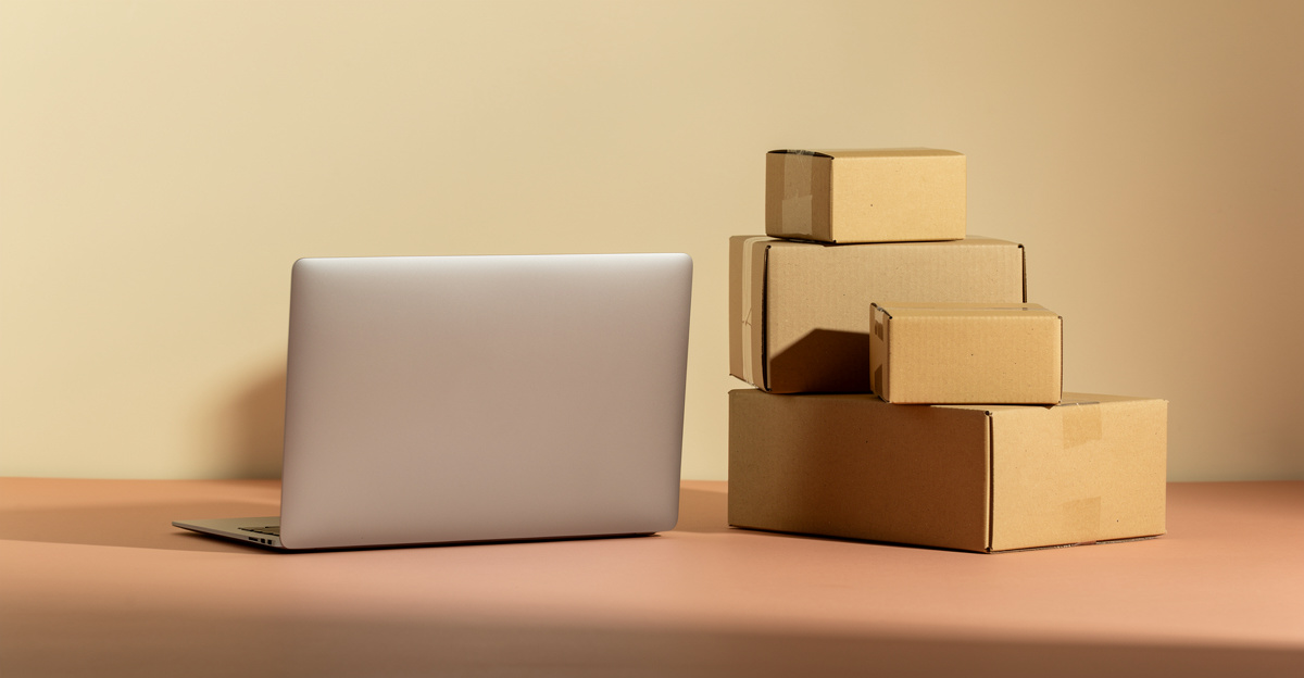 A closed laptop beside a small stack of kraft boxes on a warm cream backdrop, representing an online store operated from anywhere